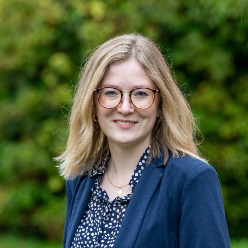Rebecca Marxcord lächelt in die Kamera. SIe hat ein dunkelblaues Jacket an, eine weiss/dunkelblaue Bluse an und eine Brille.
